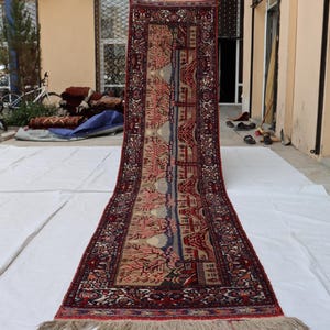 May include: A long, rectangular rug with a detailed pattern. The rug features a central design with red, blue, and beige hues, framed by a dark red border. The rug has a fringed edge.