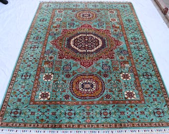 May include: A rectangular, hand-knotted rug with a central medallion design. The rug features a teal background with intricate patterns in red, brown, and cream. The border is a darker teal with floral motifs, and the rug has a fringed edge.