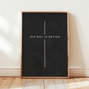 May include: A rectangular wall art print in a light wood frame featuring a black background with a thin white vertical line forming a cross shape. The text HIS WAY IS BETTER is printed in white capital letters across the center of the cross.