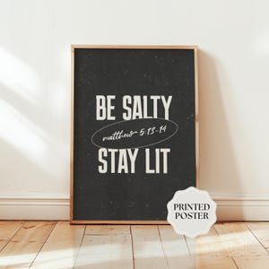 May include: A rectangular poster in a light wood frame featuring a dark charcoal background with white text that reads BE SALTY, followed by the script text matthew 5:13-14 inside an oval, and the text STAY LIT at the bottom.
