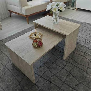 Pode incluir: Uma mesa de centro de pedra clara de dois níveis. O nível superior contém um vaso de vidro transparente com flores brancas. O nível inferior exibe objetos decorativos, incluindo uma bandeja dourada em forma de folha e um prato pequeno de fruta.