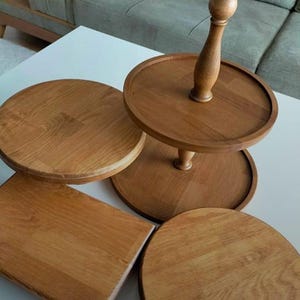 May include: A two-tiered wooden serving stand with a central spindle, accompanied by a separate round wooden tray and a rectangular wooden board. All items feature a natural light brown wood grain finish and smooth, circular or rectangular edges.