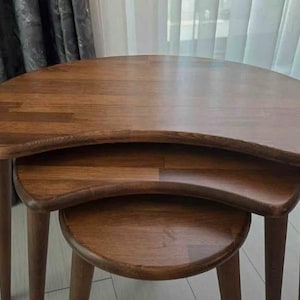 以下が含まれることがあります： ミディアムブラウンの木材で作られた3つの入れ子式テーブルのセット。各テーブルは滑らかな腎臓型の天板と3本の先細りの脚が特徴です。テーブルは積み重ねられており、全体に一貫した木目調の仕上げが見られます。
