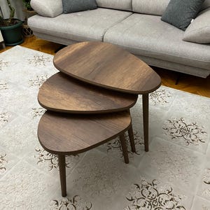 Pode incluir: Três mesas de madeira marrom aninhadas com uma forma arredondada e orgânica. As mesas estão dispostas em uma pilha, com a mesa maior na parte inferior e a mesa menor na parte superior.
