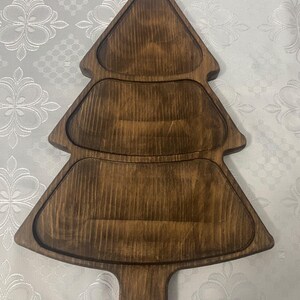 May include: A dark brown wooden serving tray shaped like a Christmas tree. The tray features three distinct recessed sections for food, with a smooth finish that highlights the natural vertical wood grain patterns throughout the surface.