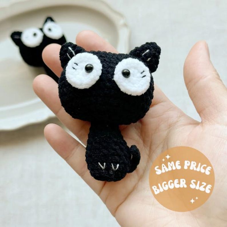 May include: A handmade black crochet cat with large white eyes and a small tail. The cat is held in a hand, with another cat in the background. A sticker on the right says "SAME PRICE BIGGER SIZE".