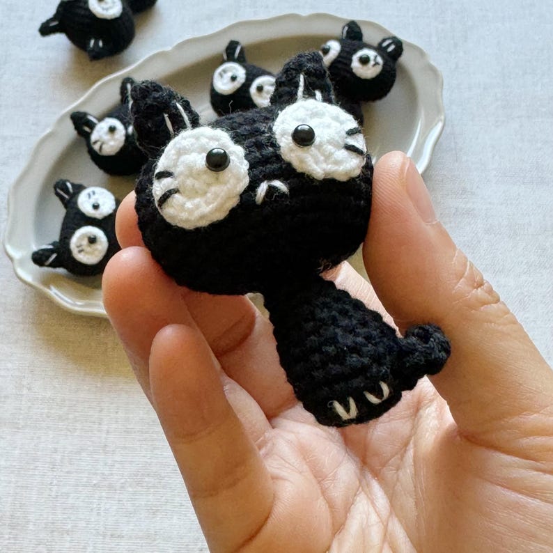 May include: A black and white crocheted cat with large white eyes and a small tail is held in a hand. Several similar cat figures are arranged on a white plate. The cats are handmade and have a whimsical appearance.