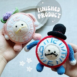 May include: Two crocheted plush toys. One is pink and white with a lion-like face and a flower crown. The other is a clock-faced character with a black hat, mustache, and blue arms and legs. The text "FINISHED PRODUCT" is in the background.
