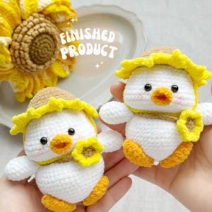 May include: Two handmade crochet ducklings with sunflower hats and accents. The ducklings are white with yellow feet, beaks, and sunflower details. The text "FINISHED PRODUCT" is visible in the background. These are likely handmade toys or decorative items.