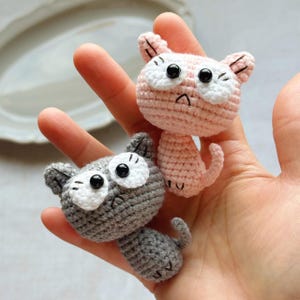 May include: Two handmade crochet cat figurines. One is light pink, the other is gray. Both have white eye accents and black pupils. The cats have small ears and curled tails. The figurines are held in a person's hand.