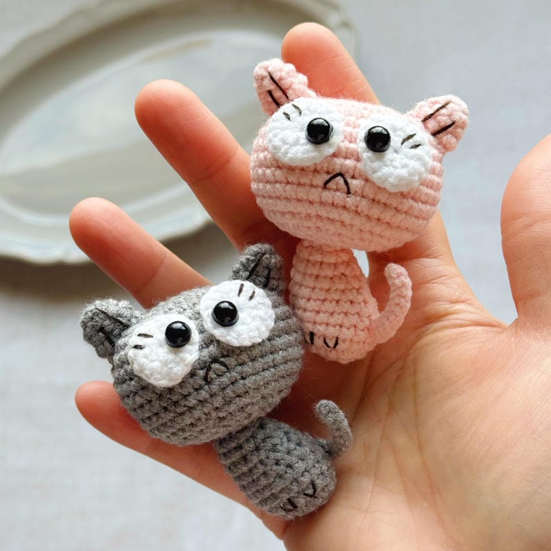 May include: Two handmade crochet cat figurines. One is light pink, the other is gray. Both have white eye accents and black pupils. The cats have small ears and curled tails. The figurines are held in a person's hand.