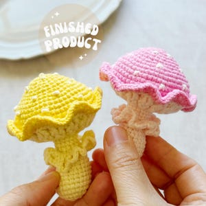May include: Two handmade crochet mushroom figures. One is yellow with white spots, the other is pink with white spots. The mushrooms have ruffled edges and are held in a person's hands. The text "FINISHED PRODUCT" is in a circle above the mushrooms.