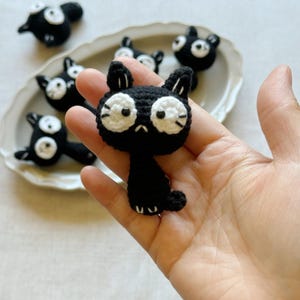 May include: Handmade black crochet cat with white eyes and whiskers. Several other similar cat figures are arranged on a white oval plate. The cats have a sad expression. The cat is held in a hand.