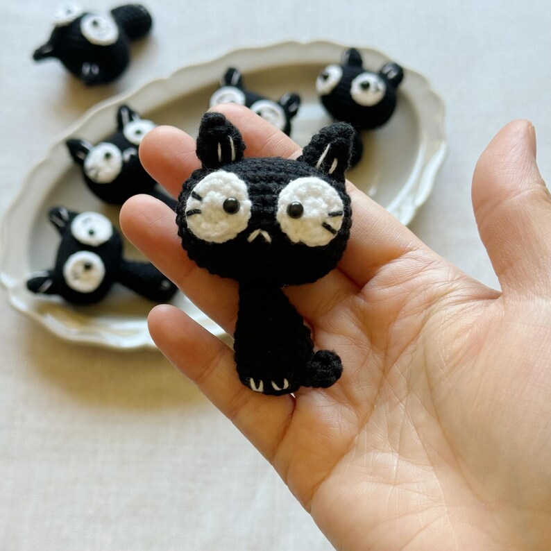 May include: Handmade black crochet cat with white eyes and whiskers. Several other similar cat figures are arranged on a white oval plate. The cats have a sad expression. The cat is held in a hand.