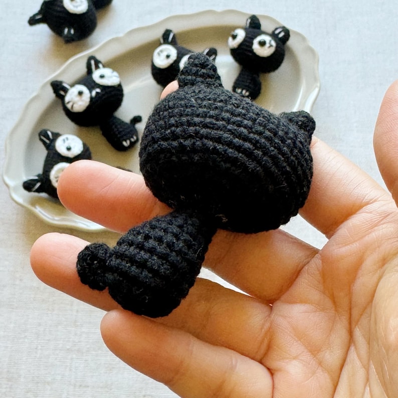 May include: Handmade black crochet cat with white eyes and a white face. Several other similar crochet cats are on a white plate. The cats have small black ears and tails. The cat is held in a hand.