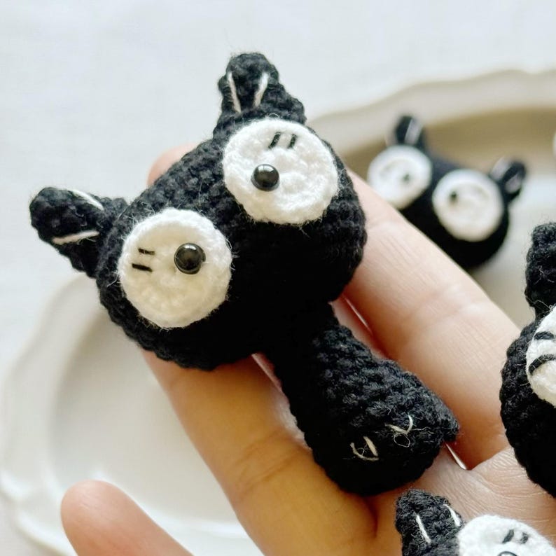 May include: Crocheted black cat with white eyes and accents. The cat has a round head, pointed ears, and a long body. The cat's eyes are large and round, with black pupils. The cat is handmade and has a whimsical appearance.