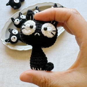 May include: A black crochet cat with large white eyes and a small, downturned mouth is held in a hand. Several similar cat figures are arranged on a white plate in the background. The cat has small ears and a curled tail.