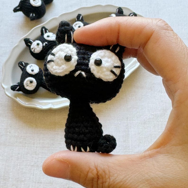 May include: A black crochet cat with large white eyes and a small, downturned mouth is held in a hand. Several similar cat figures are arranged on a white plate in the background. The cat has small ears and a curled tail.