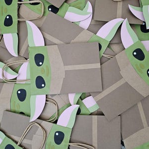 Może przedstawiać: Kolekcja brązowych papierowych torebek na prezenty z zielonymi i r&oacute;żowymi twarzami postaci. Torby mają brązowe uchwyty i są ozdobione dużymi czarnymi oczami i spiczastymi uszami. Torby są ułożone w stosie.
