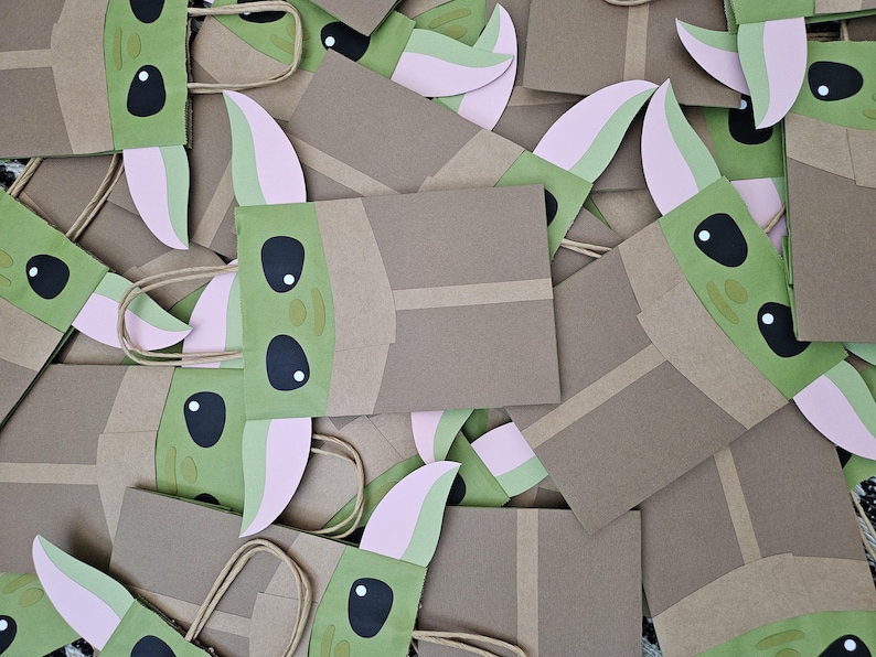 Może przedstawiać: Kolekcja brązowych papierowych torebek na prezenty z zielonymi i r&oacute;żowymi twarzami postaci. Torby mają brązowe uchwyty i są ozdobione dużymi czarnymi oczami i spiczastymi uszami. Torby są ułożone w stosie.