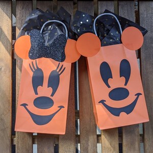 May include: Two orange Halloween treat bags with black Mickey Mouse faces. The bags have black tissue paper peeking out of the top.