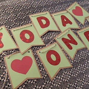 Może przedstawiać: Baner z napisem "YOU ARE ONE" w czerwonych literach i sercami na jasnozielonych proporczykach. Proporczyki mają brązową obw&oacute;dkę i są połączone sznurkiem. Tło to czarno-biała wzorzysta tkanina.