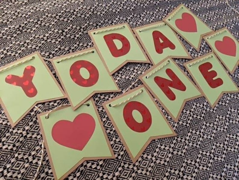 Może przedstawiać: Baner z napisem "YOU ARE ONE" w czerwonych literach i sercami na jasnozielonych proporczykach. Proporczyki mają brązową obw&oacute;dkę i są połączone sznurkiem. Tło to czarno-biała wzorzysta tkanina.