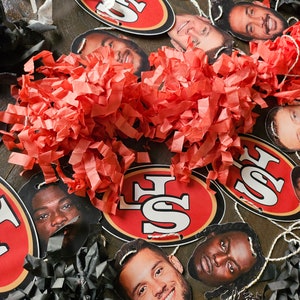 Può includere: Un set di pompon in carta rossa e nera e una ghirlanda di ritagli di carta con i volti di giocatori di football e il logo dei San Francisco 49ers.