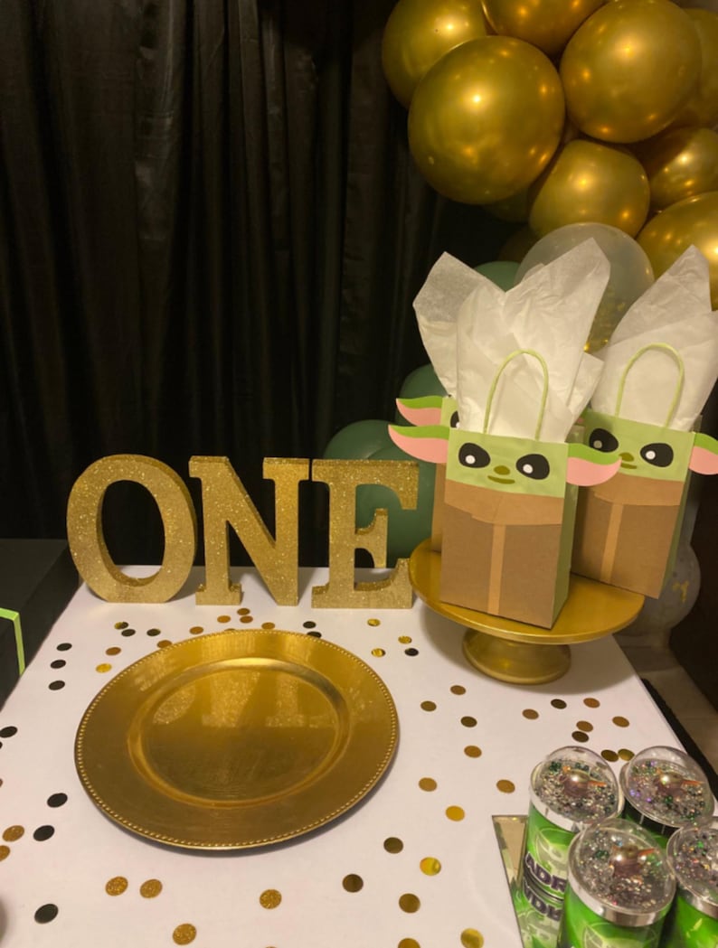 Może przedstawiać: Scena imprezy z złotymi balonami, złotym talerzem i słowem "ONE" w złotych brokatowych literach. Dwie torby na prezenty z motywem Grogu stoją na złotym stojaku na ciasto. Złote konfetti zdobi biały obrus.