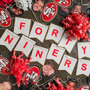 Può includere: Uno striscione rosso, bianco e oro con le parole "FORTY NINERS" in lettere rosse. Lo striscione è decorato con carta velina rossa e bianca e decorazioni a tema calcistico rosse e dorate.