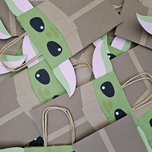 Może przedstawiać: Stos brązowych papierowych torebek na prezenty ozdobionych zieloną i r&oacute;żową twarzą postaci. Torby mają brązowe papierowe uchwyty i są ozdobione wyciętymi detalami. Postać ma duże czarne oczy i spiczaste uszy.