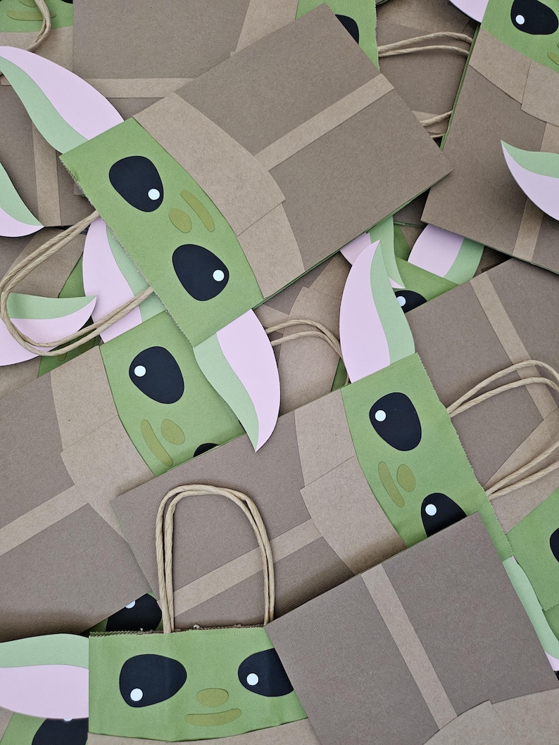 Może przedstawiać: Stos brązowych papierowych torebek na prezenty ozdobionych zieloną i r&oacute;żową twarzą postaci. Torby mają brązowe papierowe uchwyty i są ozdobione wyciętymi detalami. Postać ma duże czarne oczy i spiczaste uszy.