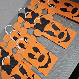 May include: Orange paper gift bags with black Mickey Mouse faces and black glitter bows. The bags have white handles and are perfect for Halloween treats.