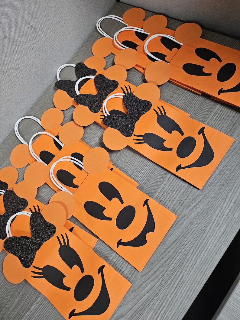 May include: Orange paper gift bags with black Mickey Mouse faces and black glitter bows. The bags have white handles and are perfect for Halloween treats.