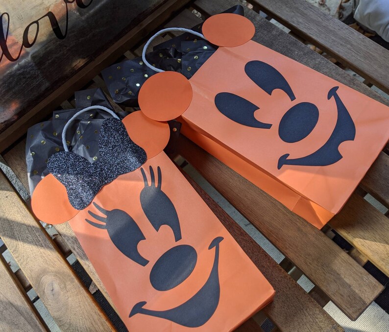 May include: Two orange Halloween treat bags with black Mickey Mouse and Minnie Mouse faces. The bags have black glitter accents and white string handles.