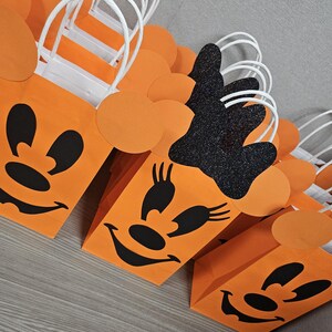 May include: Orange and black Halloween treat bags with a Minnie Mouse design. The bags have white handles and are decorated with black eyes, a nose, and a smile.