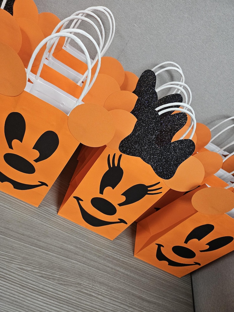 May include: Orange and black Halloween treat bags with a Minnie Mouse design. The bags have white handles and are decorated with black eyes, a nose, and a smile.