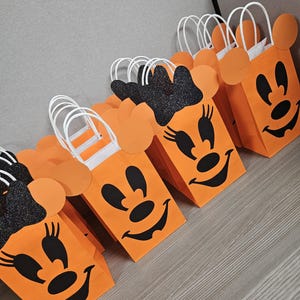 May include: Orange and black Halloween treat bags with a Minnie Mouse design. The bags have white handles and black glitter on the ears.