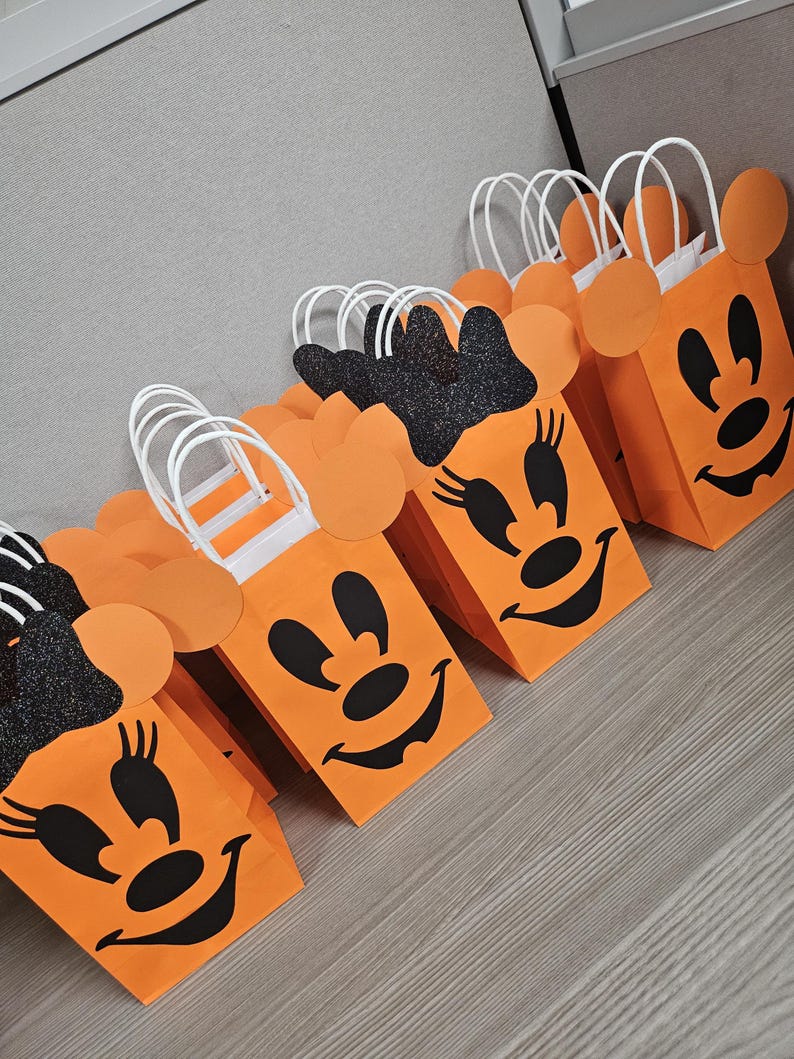 May include: Orange and black Halloween treat bags with a Minnie Mouse design. The bags have white handles and black glitter on the ears.