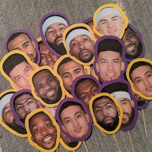 Può includere: Un set di 15 topper per cupcake viola e gialli con immagini di giocatori di basket. I topper sono realizzati in cartoncino e hanno un bastoncino attaccato sul retro.