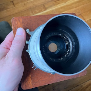 Op de afbeelding: Een vintage cameralens met een zwarte lenscilinder en een zilveren diafragmaregelring. De lens is gemonteerd op een houten basis. De tekst "G.P. GOERZ AM. OPT. CO. DAGOR 12 IN F:6" is zichtbaar op de lens.