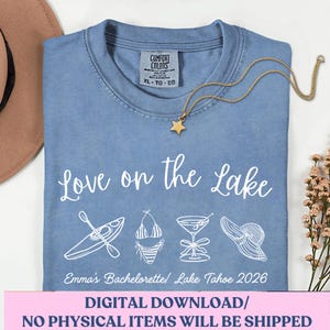 May include: A light blue t-shirt with the words "Love on the Lake" and lake-themed graphics. A gold necklace with a star pendant rests on the shirt. The text "Emma's Bachelorette/ Lake Tahoe 2026" is also visible.