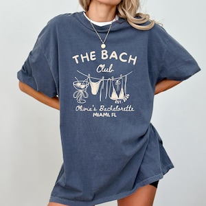 Peut inclure: Un t-shirt bleu fonc&eacute; avec l'inscription "THE BACH Club" et "Olivia's Bachelorette MIAMI, FL" en blanc, dans un style vintage. Le motif comprend un cocktail, des maillots de bain et l'ann&eacute;e "2026".