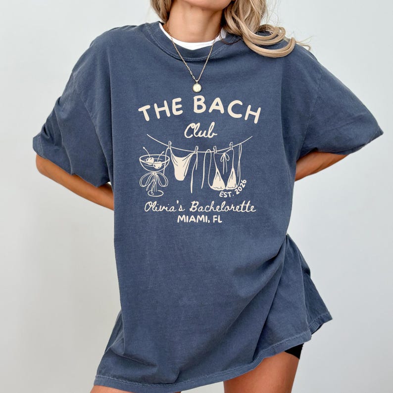 Peut inclure: Un t-shirt bleu fonc&eacute; avec l'inscription "THE BACH Club" et "Olivia's Bachelorette MIAMI, FL" en blanc, dans un style vintage. Le motif comprend un cocktail, des maillots de bain et l'ann&eacute;e "2026".