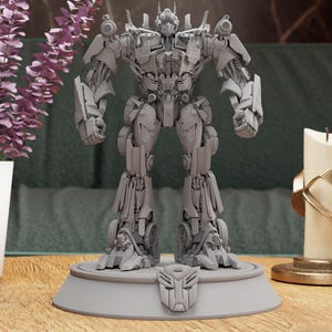 Pode incluir: Um modelo de plástico cinza e detalhado do Optimus Prime, um robô da franquia Transformers, em pé sobre uma base cinza com o símbolo dos Autobots. O modelo está em posição de pé com os braços ao lado do corpo.