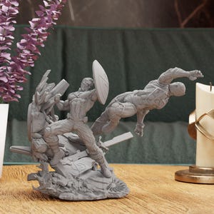 Puede incluir: Una figura impresa en 3D de color gris de Captain America y otro superhéroe en una pose dinámica. La figura está colocada sobre una base con una superficie texturizada y escombros.