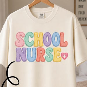 May include: An ivory short-sleeve t-shirt featuring the text SCHOOL NURSE in a multi-colored faux patchwork design with a small heart and plus sign icon. The letters are outlined in a dashed stitch pattern in shades of pink, blue, yellow, and green.