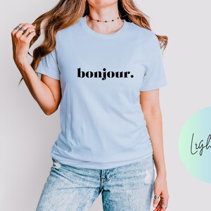 Puede incluir: Una camiseta azul claro con la palabra "bonjour." impresa en negro.