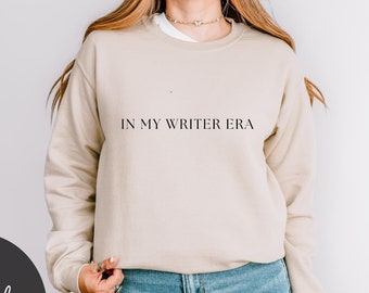 May include: A beige sweatshirt with the text "IN MY WRITER ERA" printed on the front. The sweatshirt is being worn by a person with long blonde hair. The person is wearing blue jeans.
