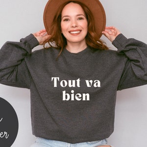 May include: A dark heather grey sweatshirt with the French phrase "Tout va bien" printed in white on the front.
